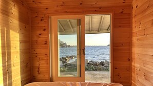 A wooden cabin room with a bed, a door leading to a rocky shoreline, and a view of the water.