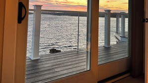 A room with a large sliding glass door leading to a balcony with a view of a sunset over water and a bridge.