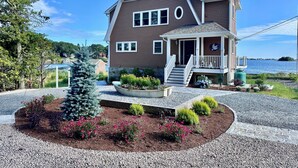 A two-story house with a porch, a small garden with flowers and a tree, and a view of the water.