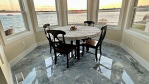 A dining area with a round table, four chairs, and a chandelier. It features large windows with a view of the water and a sunset. The floor is tiled with a marble pattern.