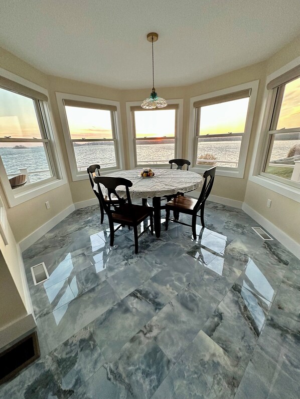 Un comedor con una mesa redonda, cuatro sillas y una araña. Cuenta con ventanas amplias que ofrecen vistas al agua y al atardecer. El piso está cubierto de baldosas con diseño de mármol.
