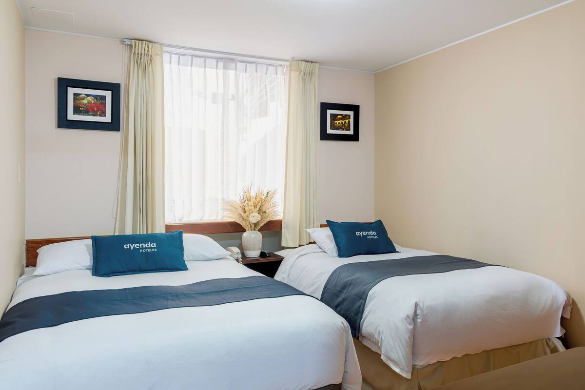 Ein Hotelzimmer mit zwei Betten, jedes mit einem Kissen mit dem Aufdruck "ayenda hotels". An den Wänden hängen zwei gerahmte Bilder, ein Fenster mit Vorhängen und ein Nachttisch mit einer Vase mit Trockenblumen.