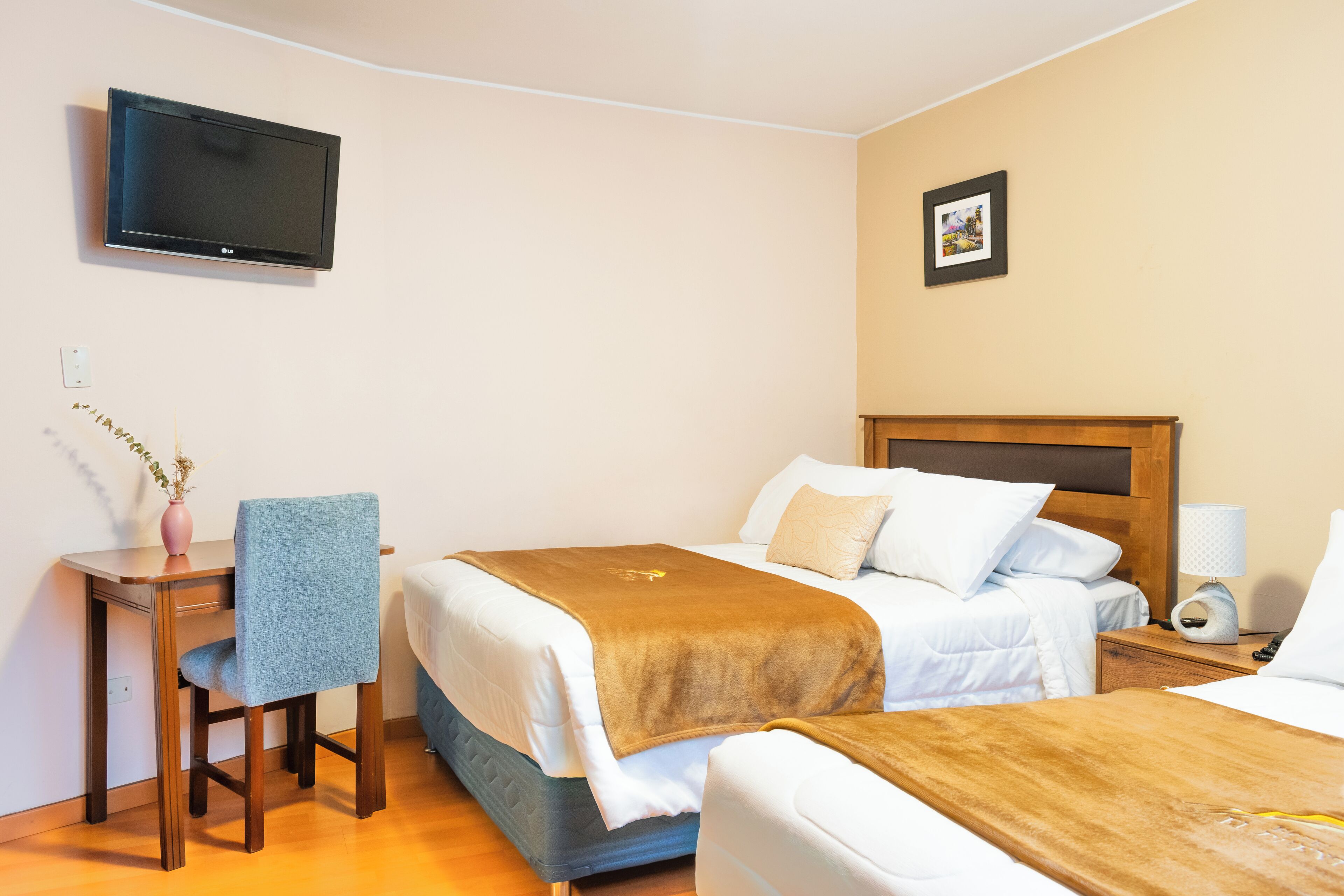 A hotel room with two beds, a TV mounted on the wall, a desk with a chair, and a small table with a vase.