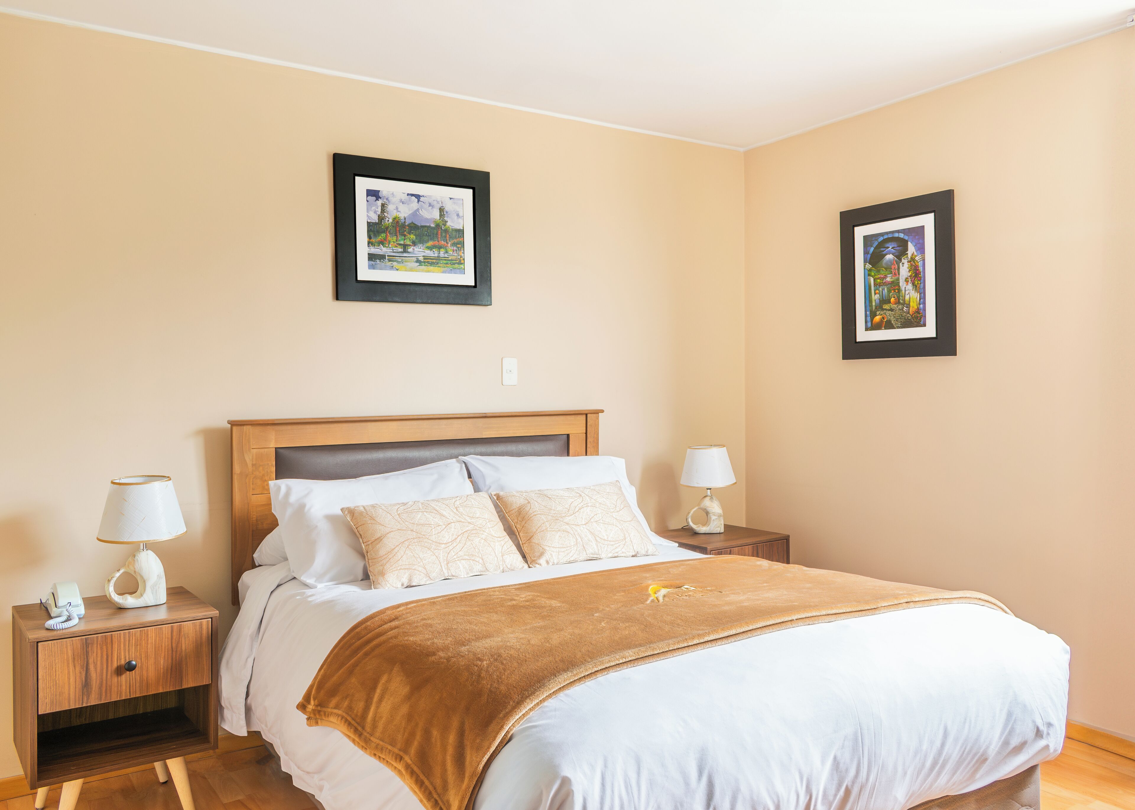 A hotel room with a bed, two bedside tables, a nightstand, and two framed pictures on the wall.