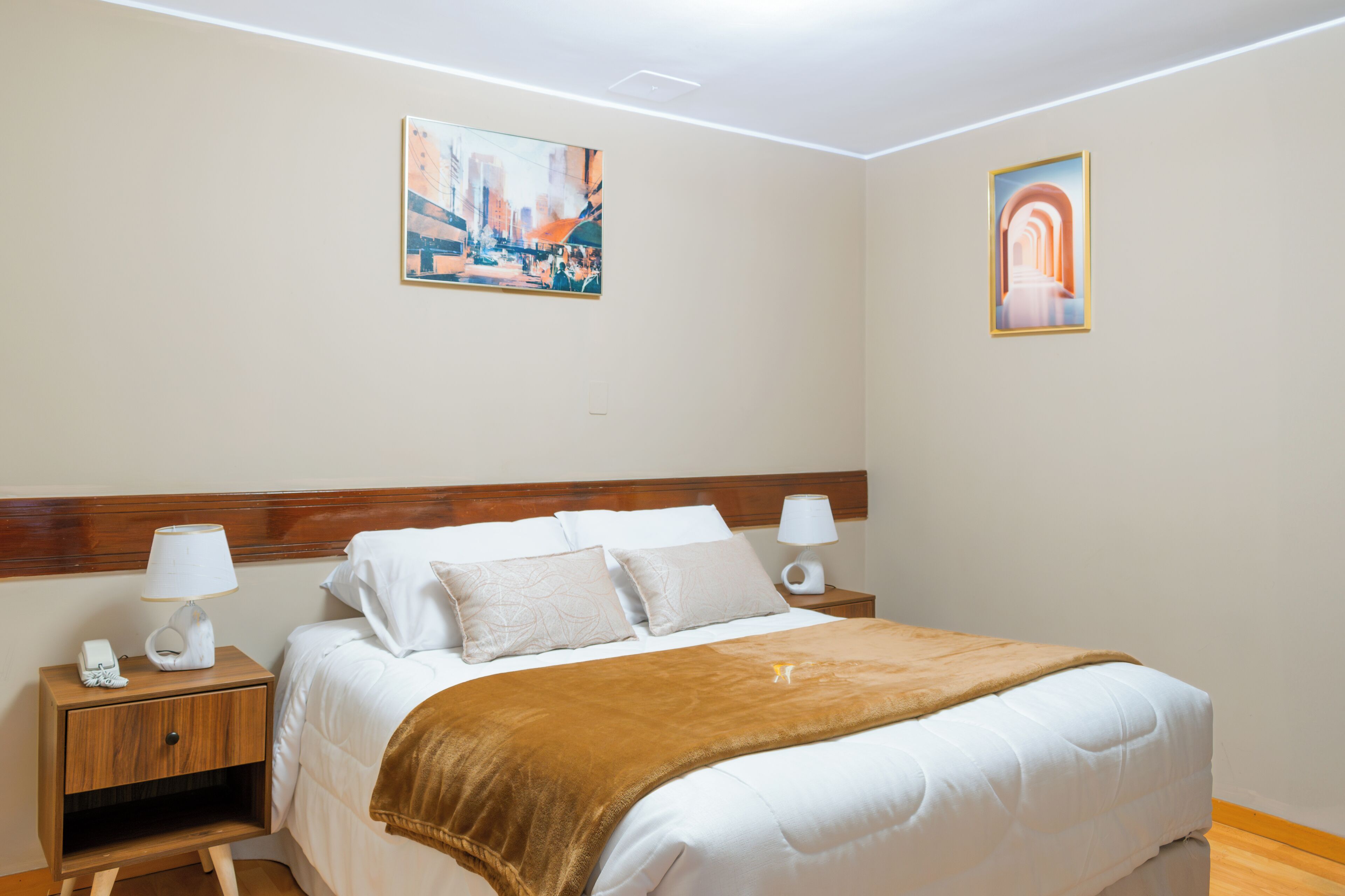 A hotel room with a large bed, two bedside tables, a nightstand, a lamp, and two framed pictures on the wall.