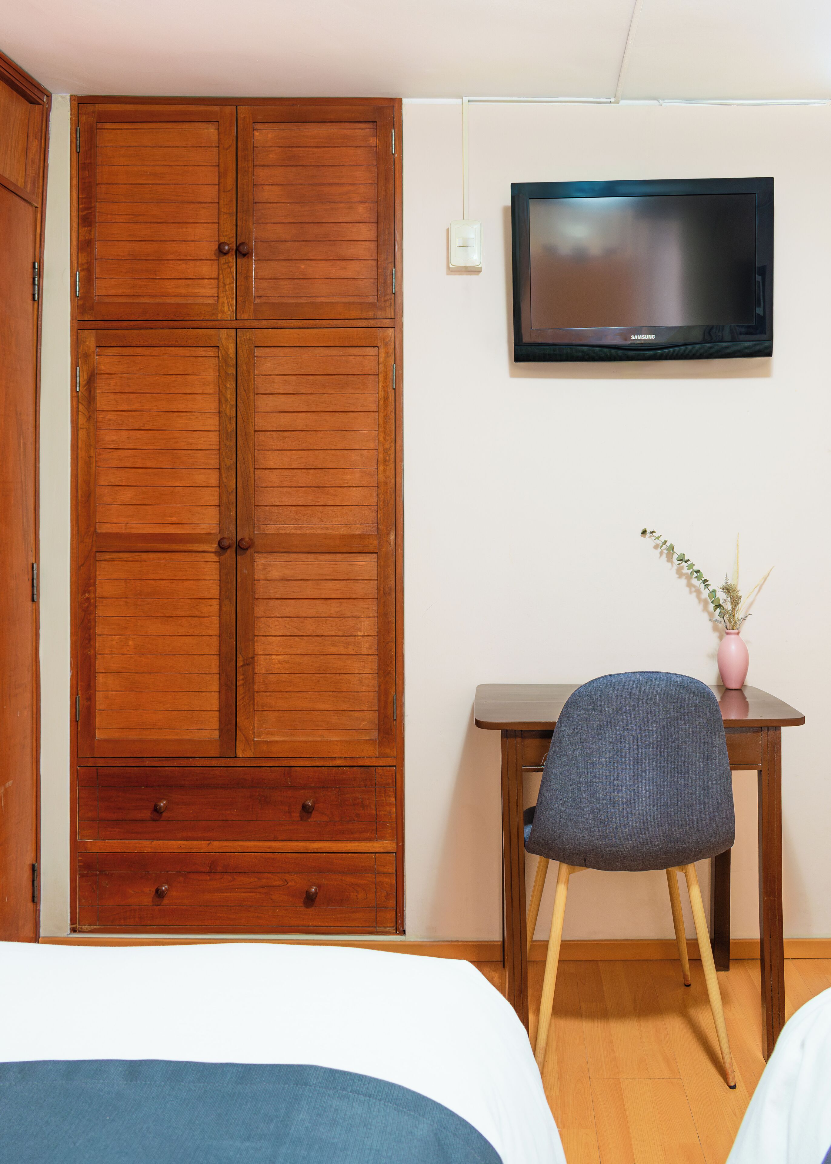 A bedroom with a wooden wardrobe, a television on the wall, a desk with a chair, and a bed.