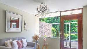 Un salon avec un canapé gris, un lustre, un tableau encadré et une vue sur un espace extérieur verdoyant, accessible par une porte vitrée.