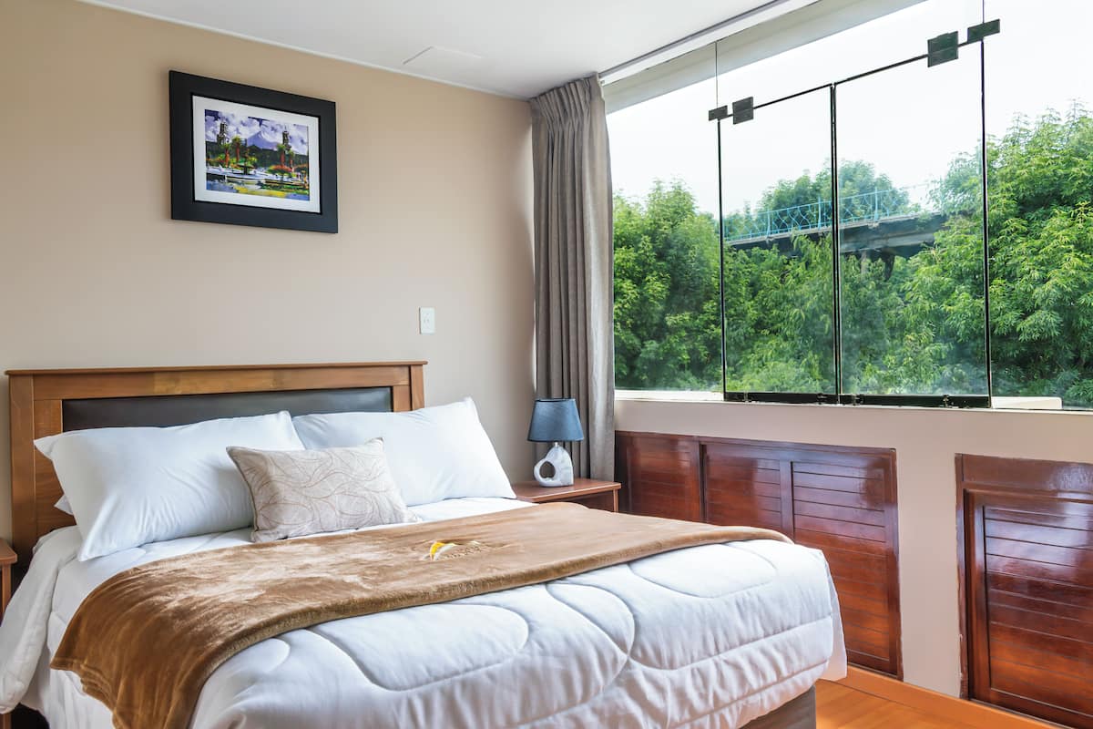 Ein Schlafzimmer mit einem großen Bett, einem Nachttisch, einer Lampe, einem gerahmten Bild an der Wand und einem Fenster mit Blick auf Grünflächen und eine Brücke.