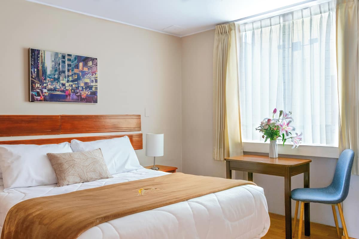 Ein Hotelzimmer mit einem großen Bett, einem Holzkopfteil, einem Nachttisch mit Lampe, einem kleinen Tisch mit Blumenvase und einem Fenster mit Vorhängen.