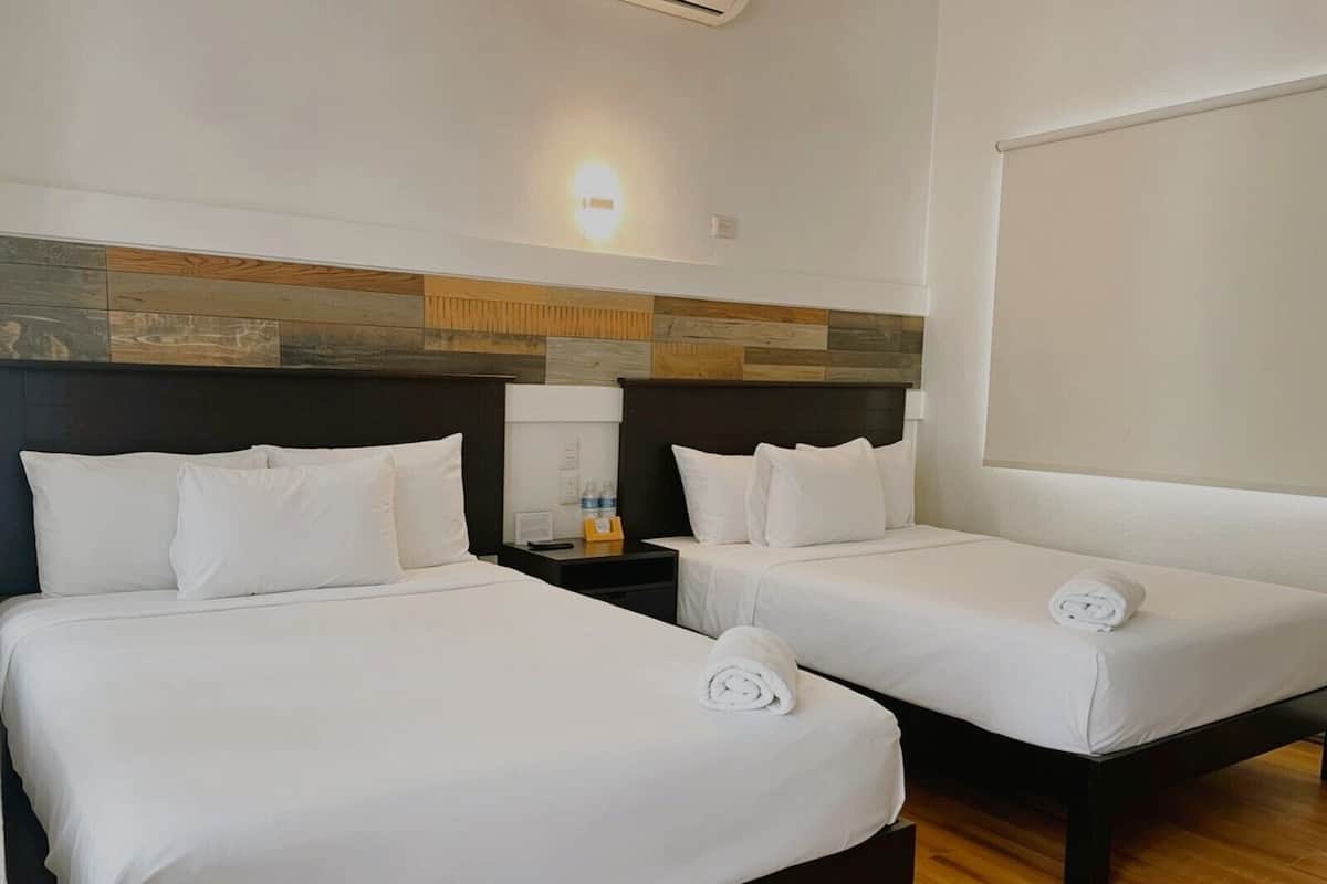 Quarto de hotel com duas camas, uma mesinha de cabeceira, uma luminária na parede e uma cabeceira de madeira.
