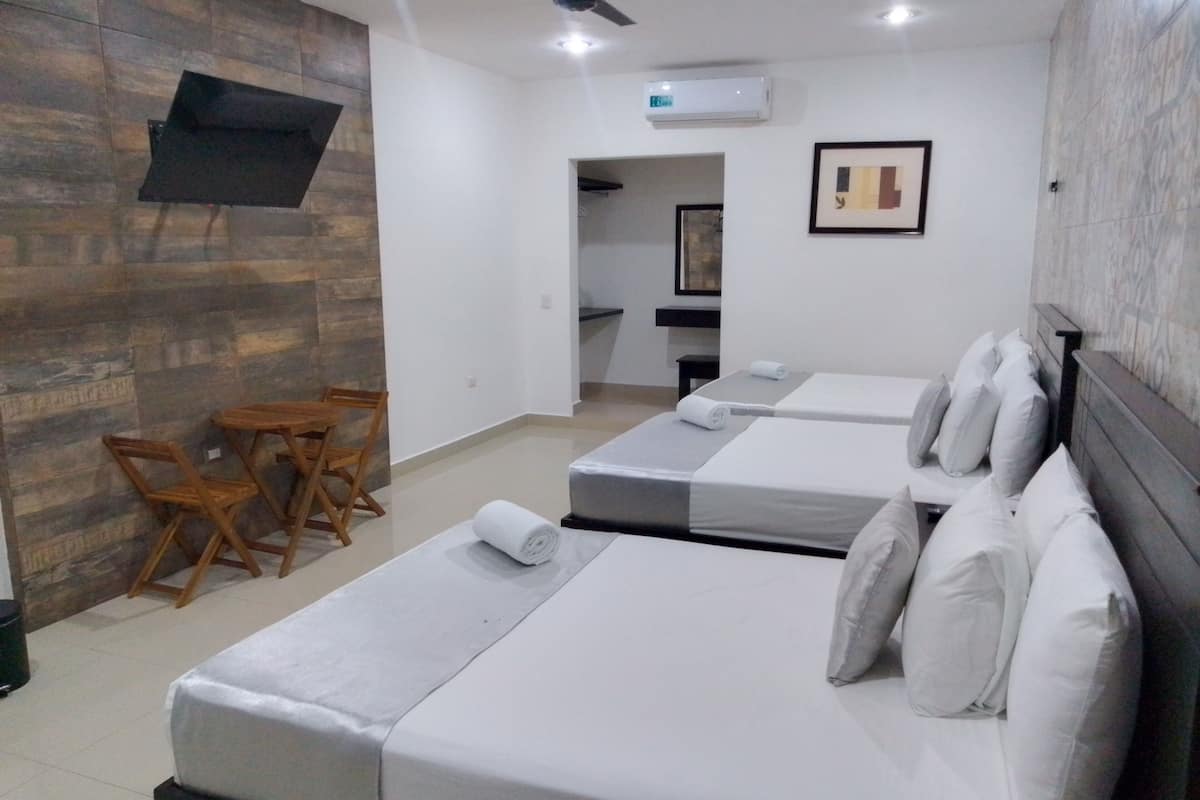 Quarto de hotel com três camas, parede de madeira, televisão de tela plana e uma pequena mesa com cadeiras.
