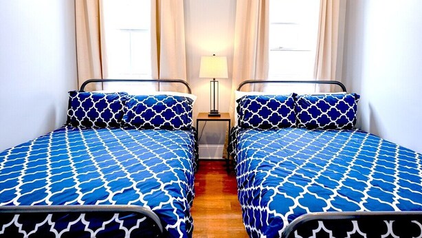 A room with two beds, each with a patterned blue and white bedspread, a bedside table with a lamp, and a window with curtains. - Governors Apartment -  2 bedroom  - Steps to FQ (New Orleans)