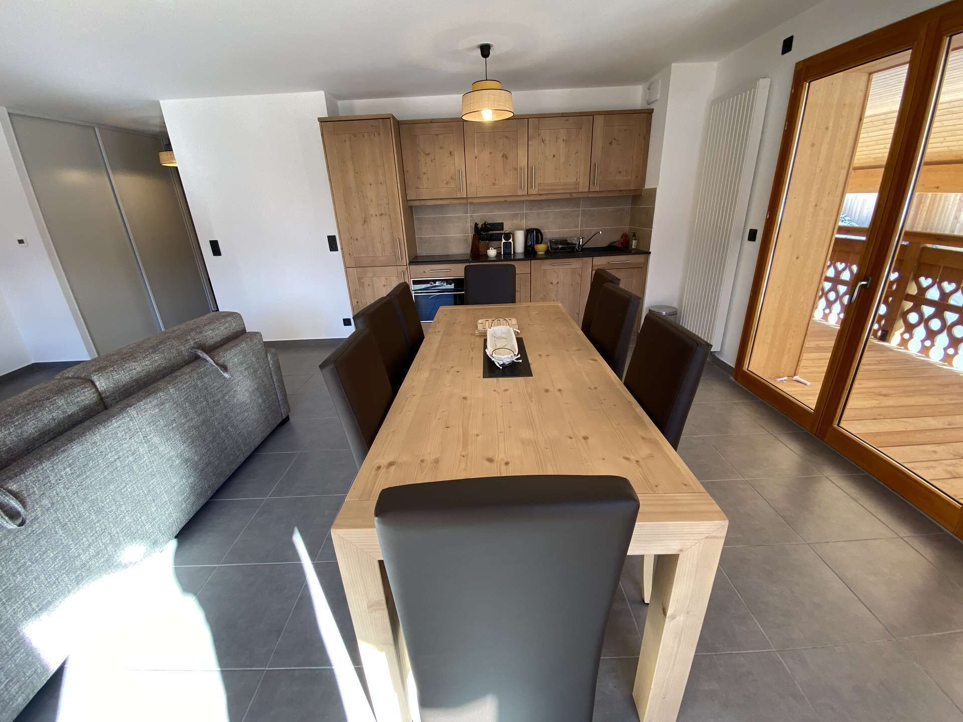 A modern kitchen with wooden cabinets, a dining table with chairs, and a grey sofa.