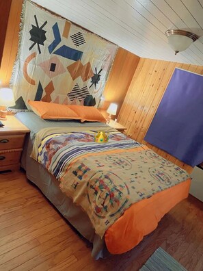 A bedroom with a wooden floor, a bed with a colorful blanket, two bedside tables with lamps, and a wooden headboard with a geometric curtain.