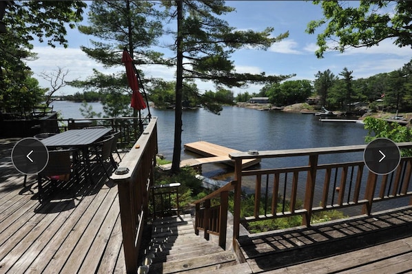 A wooden deck with a table and chairs, overlooking a calm lake with docks and boats.