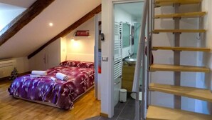 Un dormitorio con colcha roja, viga de madera en el techo, una escalera y un baño con lavabo y espejo.
