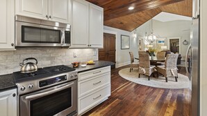 Une cuisine avec des meubles blancs, des appareils en acier inoxydable, un plafond en bois et un coin repas avec des chaises en osier.