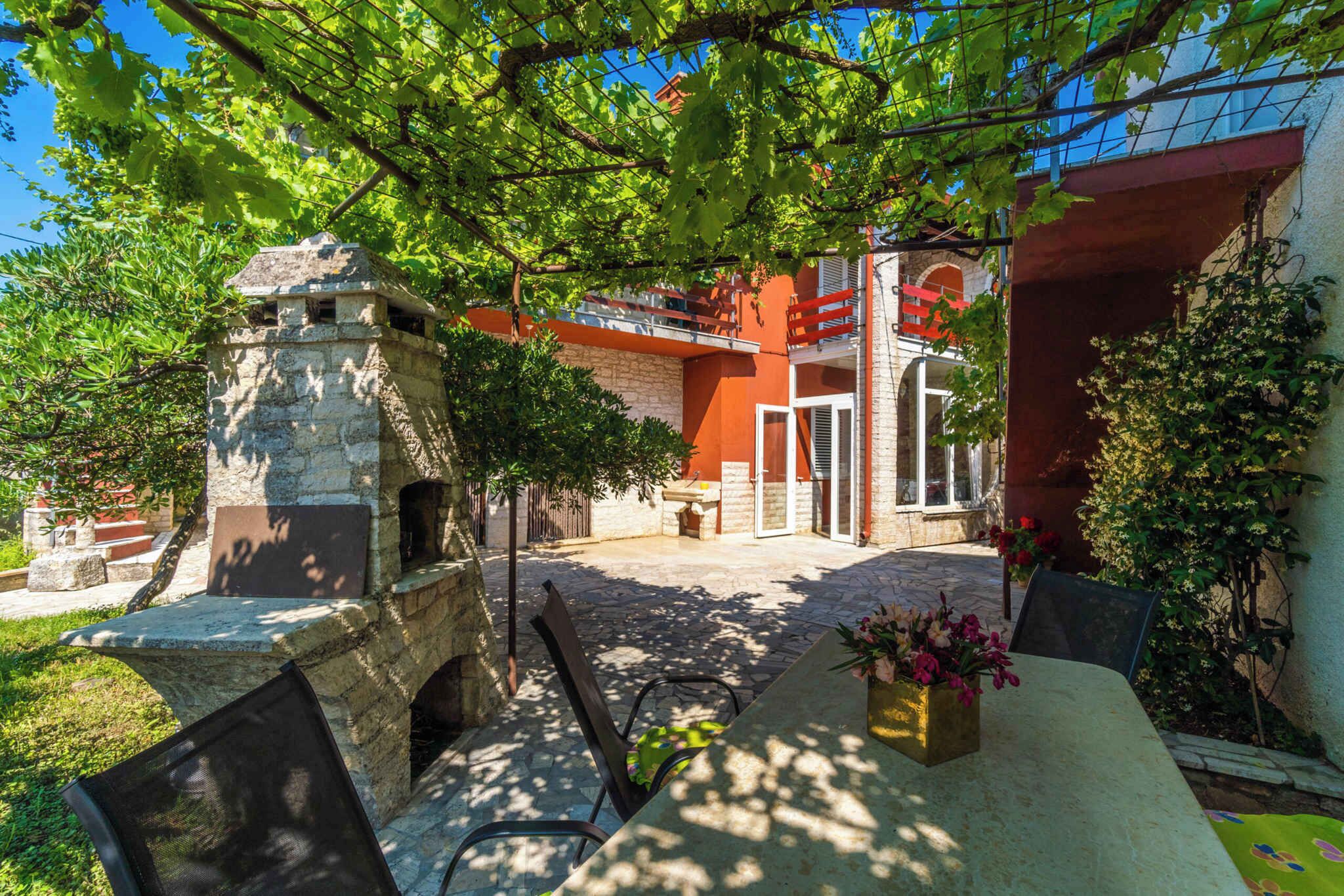 A stone structure with a table and chairs in a courtyard, surrounded by greenery and a building with red and white walls.