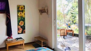 A room with a bed, a wooden bench, a small table with books, a painting, and a sliding glass door leading to an outdoor area with a bench and greenery. - Sunnybrae Garden Studio (Arcata)