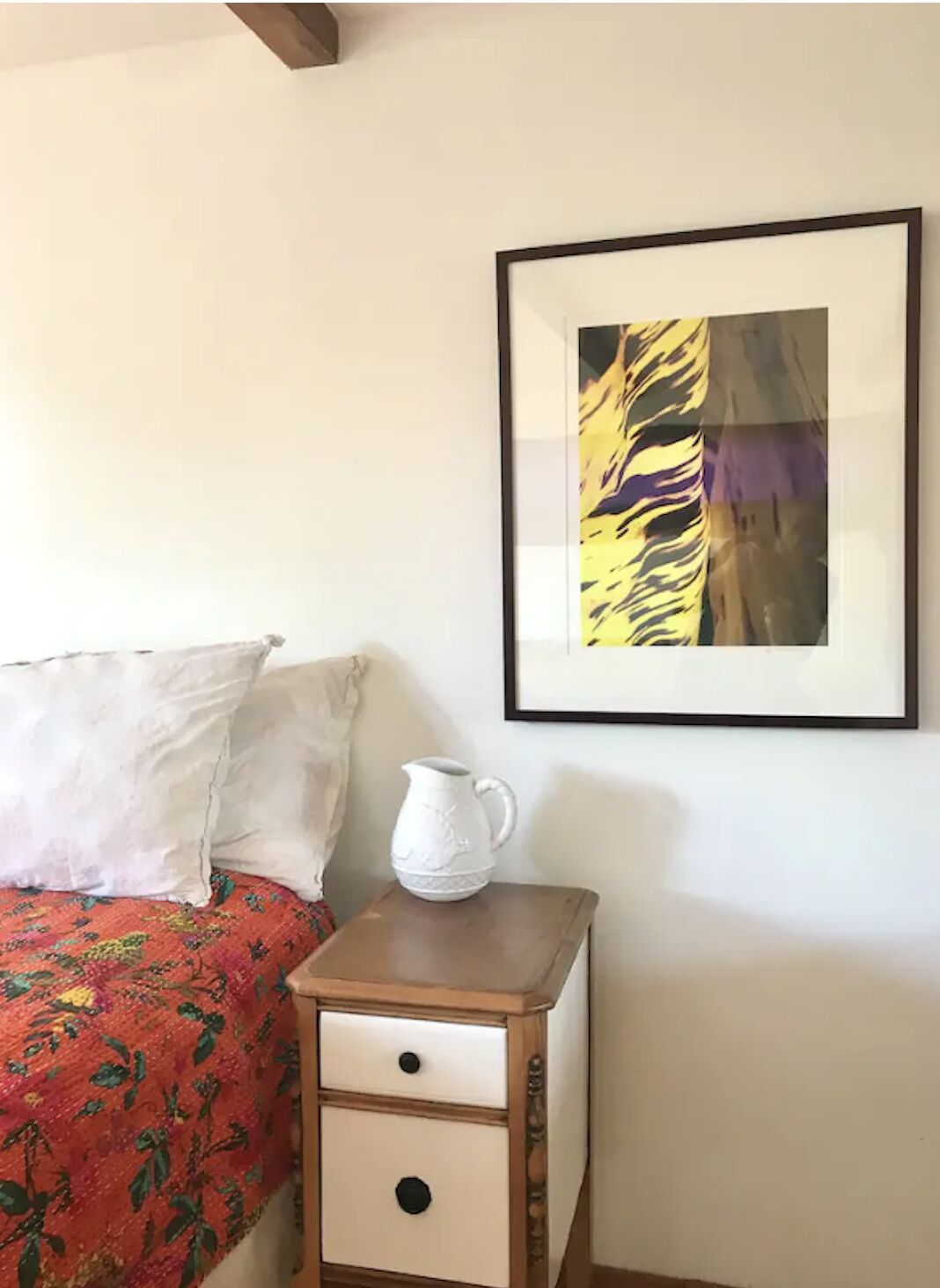 Une chambre à coucher avec un lit, des oreillers, une table de chevet, une carafe et un tableau abstrait encadré accroché au mur.