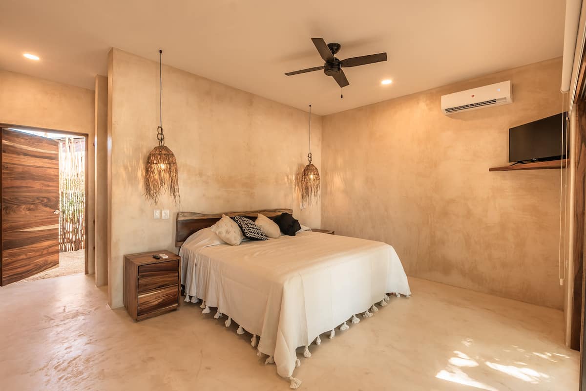 A bedroom with a bed, bedside tables, a ceiling fan, an air conditioning unit, and a TV mounted on the wall.