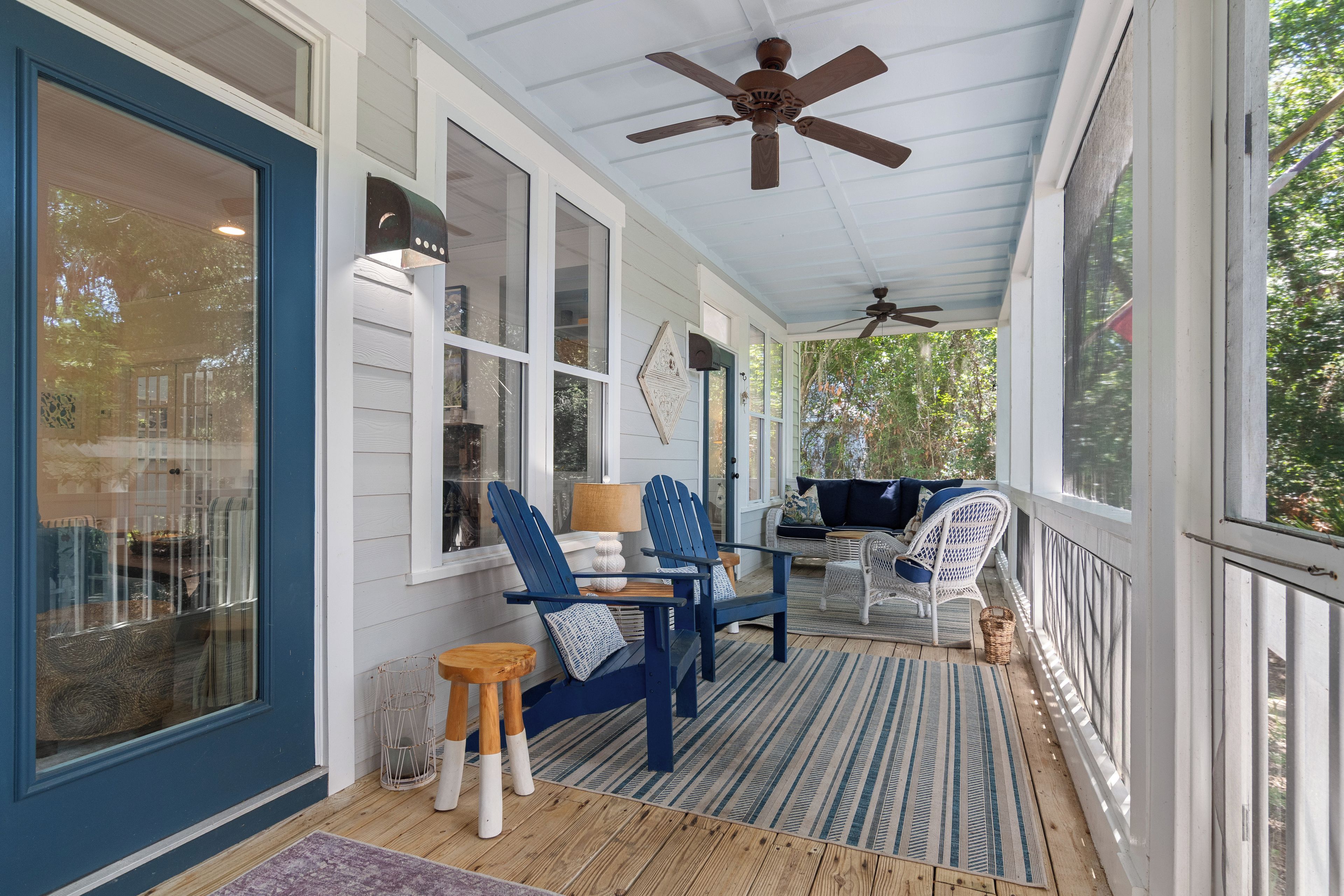 Eine überdachte Veranda mit Deckenventilator, blauen Adirondack-Stühlen, einem Tisch mit Lampe und einem Korbwäscheschrank.