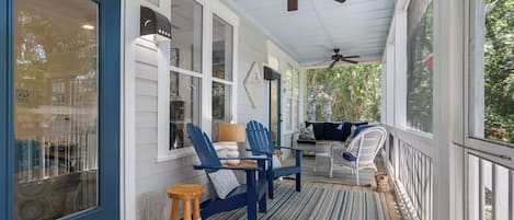 Eine überdachte Veranda mit Deckenventilator, blauen Adirondack-Stühlen, einem Tisch mit Lampe und einem Korbwäscheschrank.