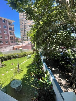 Un jardín cuidado con césped, una mesa de metal y sillas, rodeado por un muro bajo y árboles.