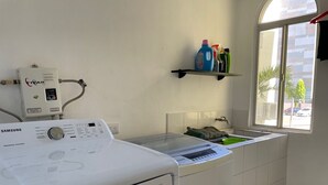 A laundry area with a Samsung washer and dryer, a small window, and a shelf with cleaning supplies. - 5 minutos Zona Rosa, Museos, Monumentos, Centros Comerciales. Casa Presidencial (San Salvador)