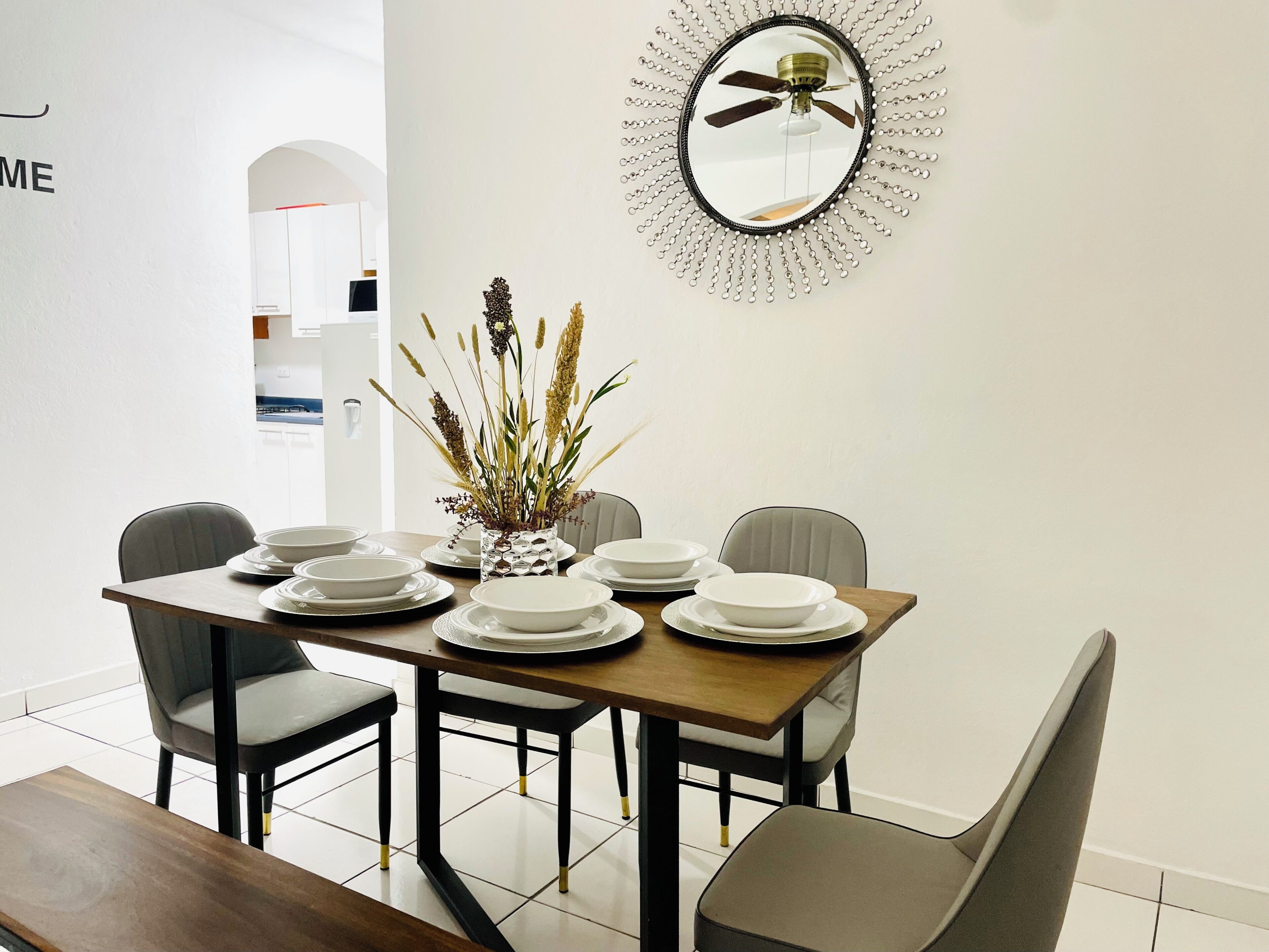 Un comedor con una mesa dispuesta para cuatro personas, sillas, un jarrón decorativo con plantas y un espejo colgado en la pared.