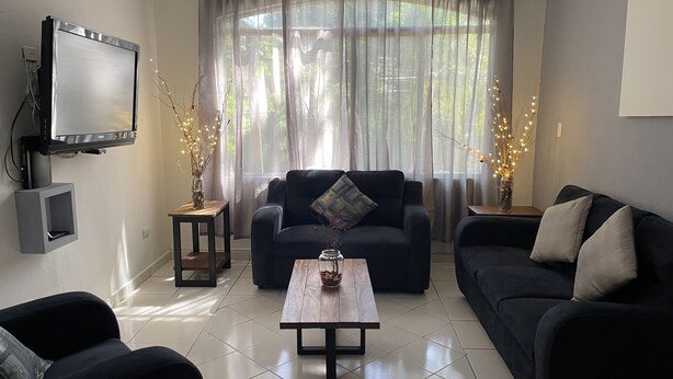 A living room with a dark sofa, a coffee table, and a TV mounted on the wall. - 5 minutos Zona Rosa, Museos, Monumentos, Centros Comerciales. Casa Presidencial (San Salvador)