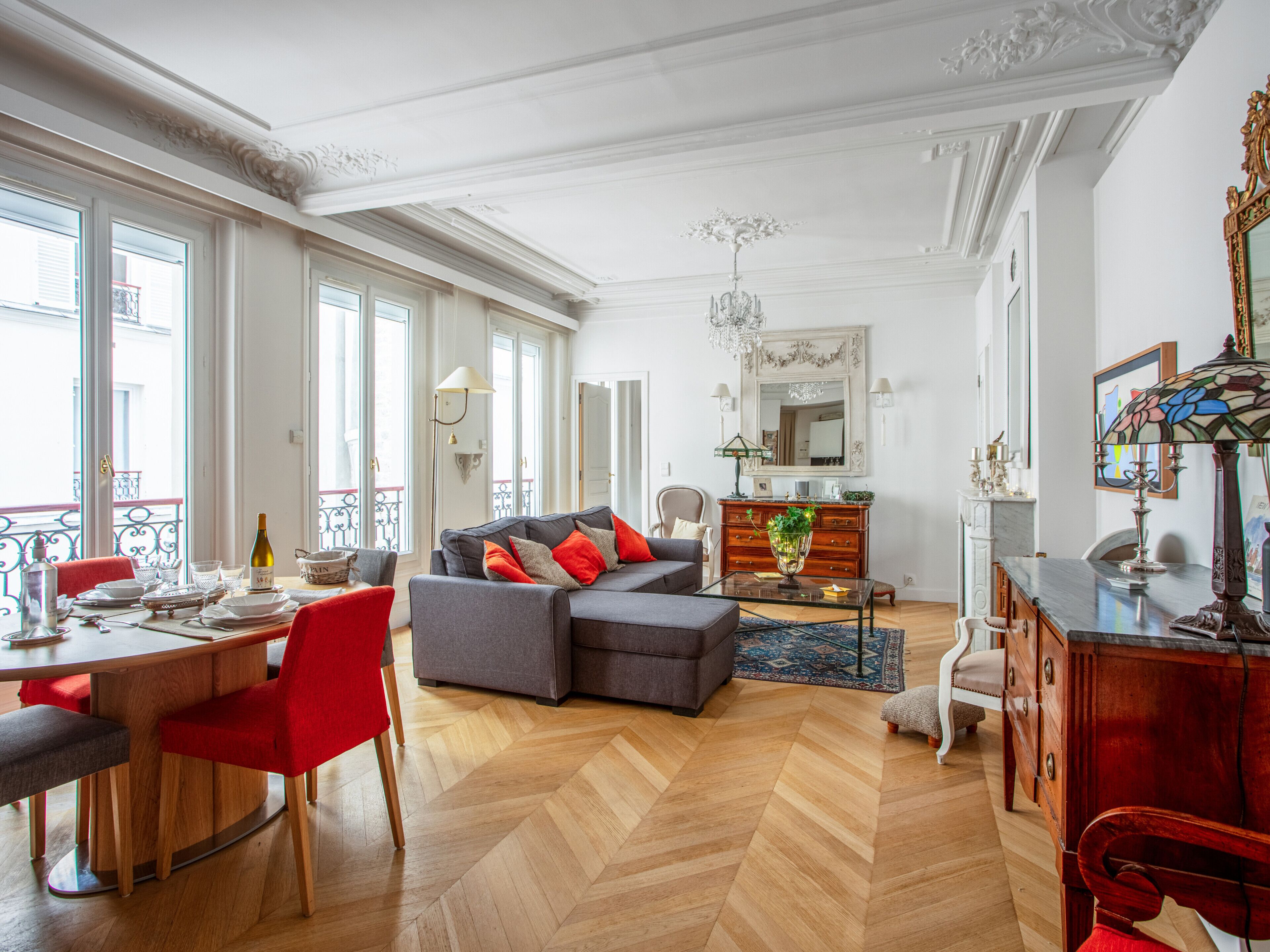A spacious living room with a herringbone wood floor, a grey sofa with red cushions, a wooden coffee table, and a dining area with a red chair and a table set for two.