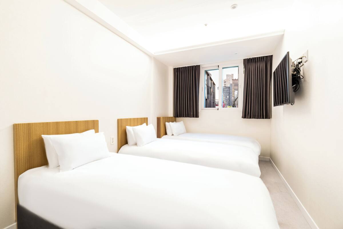 A hotel room with two beds, a large window with curtains, a flat-screen TV mounted on the wall, and a city view.