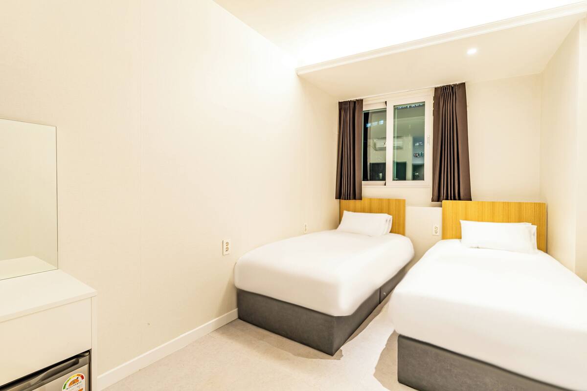 A hotel room with two single beds, a window with curtains, a small bedside table, and a wardrobe.