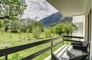 Varanda com mobiliário de vime, vista para área gramada com árvores e montanhas ao fundo.