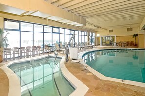 An indoor swimming pool with clear glass walls, a tiled floor, and a seating area with chairs.