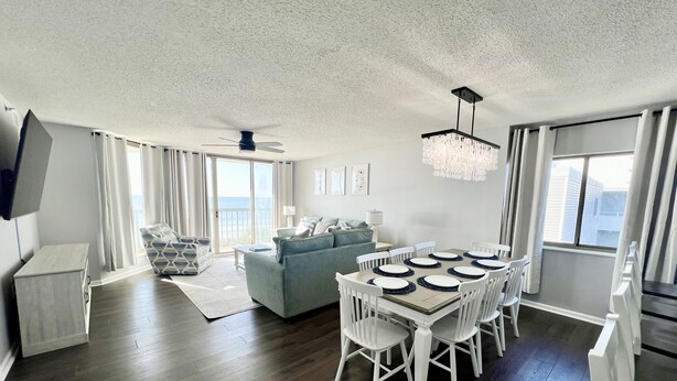 A spacious living room with a dining area, a sofa, a TV, and a chandelier. - Emerald Oasis - Oceanfront 3rd FL End Unit - 4BD/3BA - Newly Renovated Feb '24 (North Myrtle Beach)