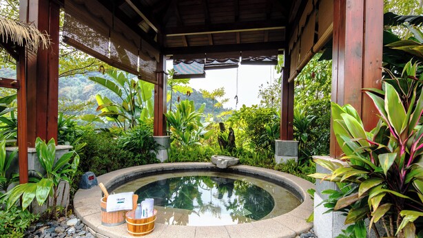 Ein friedlicher Garten mit einem runden Whirlpool, umgeben von üppiger Vegetation und einem hölzernen Pavillon.