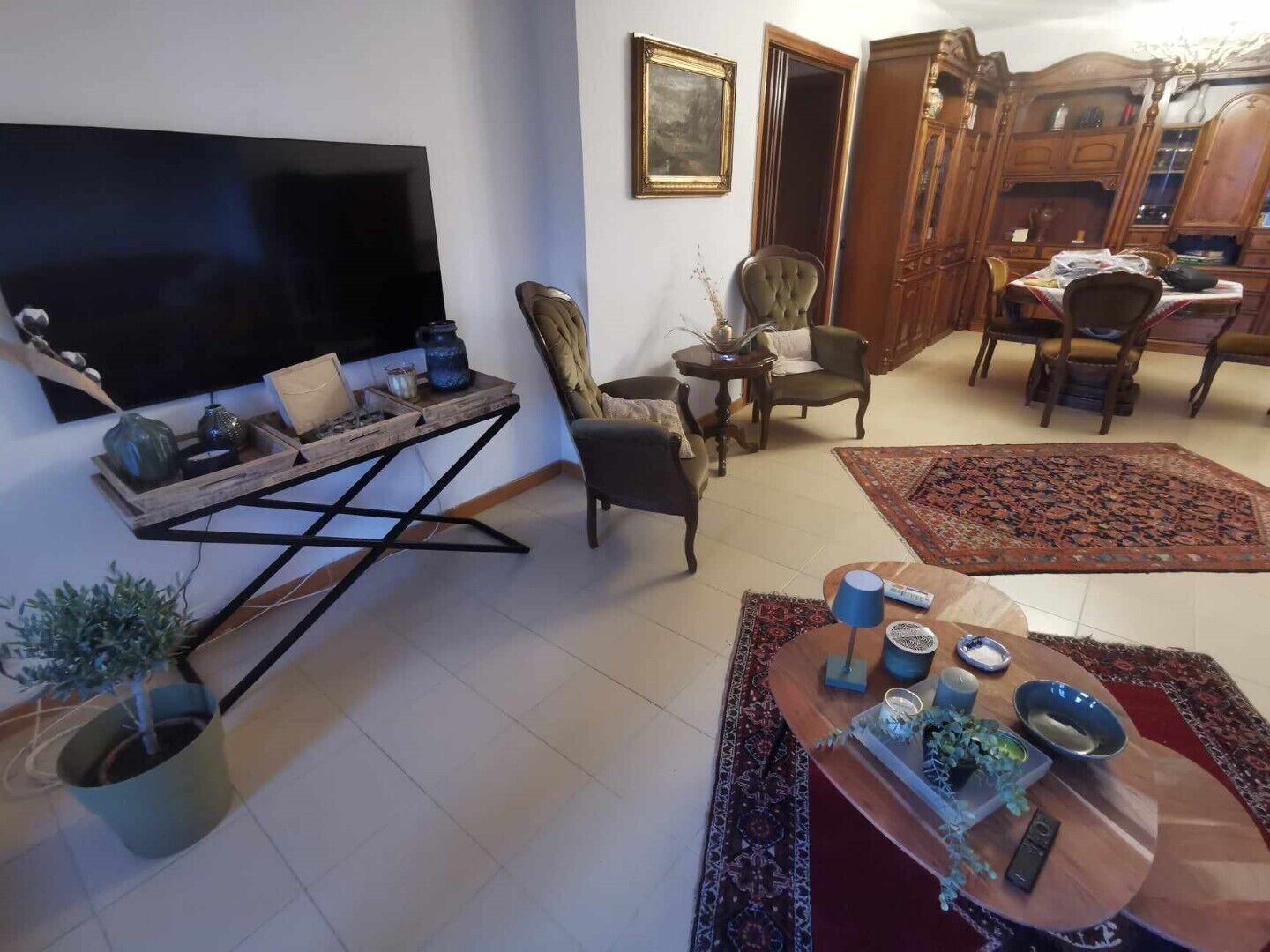 A living room with a flat-screen TV, a wooden coffee table, a patterned rug, and a dining area with chairs and a table.