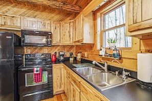 Una cocina con armarios de madera, un lavamanos de acero inoxidable, un microondas, una estufa y una ventana con vistas a árboles.