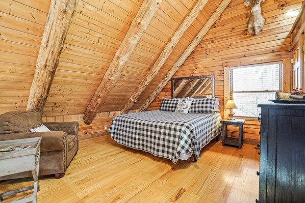 A cozy attic bedroom with a wooden ceiling, a bed with checkered bedding, a small table with a lamp, and a window with blinds.
