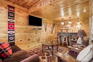 Interior de una cabaña de madera con sofá, mesa, lámpara, televisor montado y pared de piedra.