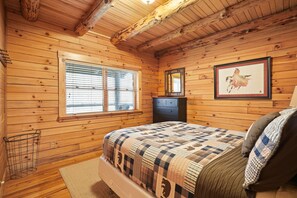 Ein Schlafzimmer einer Holzhütte mit Bett, Fenster, Kommode und einem gerahmten Bild an der Wand.