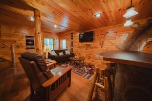 A log cabin interior with a wooden ceiling, a leather sofa, a dining table, and a mounted animal head.