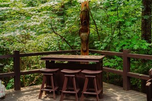 A wooden deck with a table and three stools, surrounded by a wooden railing, set in a lush green forest.