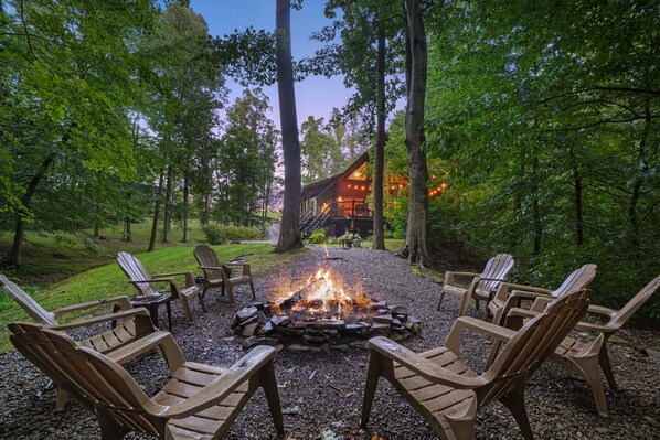 A fire pit surrounded by Adirondack chairs in a forested area with a lodge in the background.