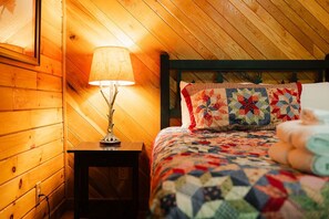 A cozy room with wooden walls, a bed with a quilt, a bedside lamp, and a framed picture on the wall.