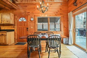A wooden cabin interior with a dining table, four chairs, a chandelier, a window with blinds, and a door with a clock. - Arrowhead Lodge | Hocking Hills, Hot Tub, 5 Acres (Logan)
