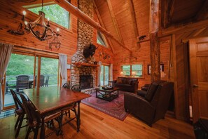 A log cabin interior with a stone fireplace, wooden dining table, leather sofa, and large windows.
