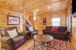 Un interior de cabaña acogedor con paredes de madera, un sillón de cuero, un sofá, una mesa de centro y una escalera.